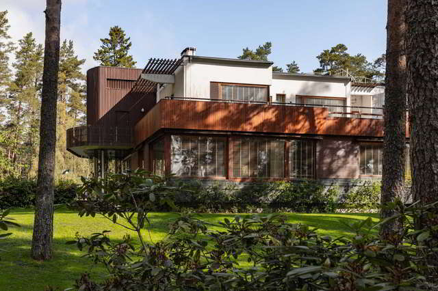 A house with large windows and wooden and white rendered facades in a park-like setting with pine trees and rhododendrons
