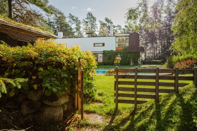 A gate in a wooden fence next to a wooden building, pool and house in the background