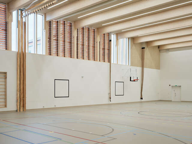 An interior photograph of a sports hall, with narrow vertical windows along the upper walls and large timber beams visible beneath the roof.
