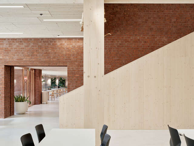 An interior photo featuring a timber wall railing in front of a brick wall. An opening on the right reveals a hallway with tables.