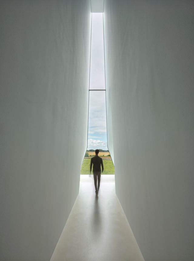 A narrow, tall corridor leads to a window overlooking an agricultural landscape. A man walking in the centre toward the window.