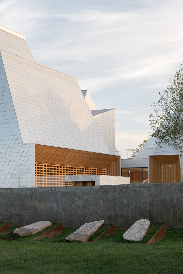 A brutalist stone wall, with a tall aluminium building behind it, featuring timber cladding at the lower level.