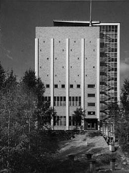 Black and white picture of a tall brick building with a glass wall stairway on the right edge of the building.