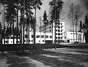 Black and white picture of a white multiple story building from between trees.