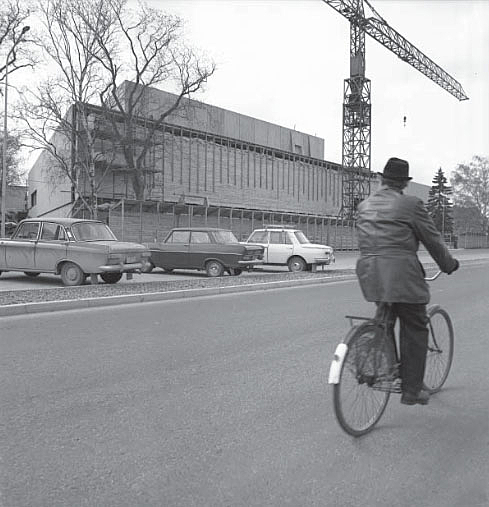 Man rides a bike next to a construction site, in front of it there are old cars.