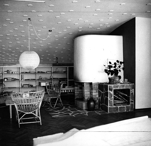 Old picture of a living room with a fireplace, bookshelves at the back of the room.