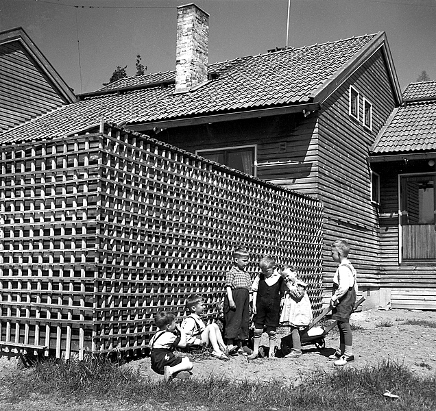 Children playing in front of a wooden fence, one boy has a wheelbarrow.