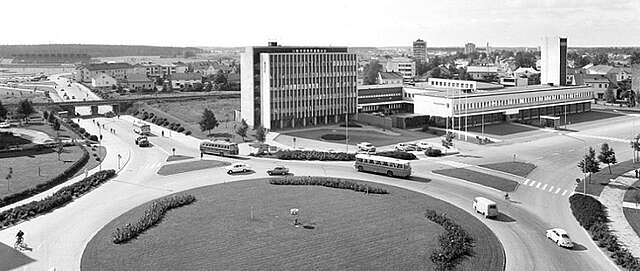 Black and white picture of a roundabout with old cars in it and commercial buildings in the background.