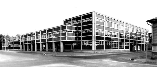 Black and white picture of the building facade. The right side has three floors while the left side has only two along with a protruding extension on the second level which is supported by pillars.