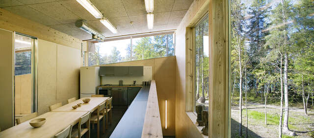 long and narrow room with wooden surfaces, a long dining table and a kitchen at the back.
