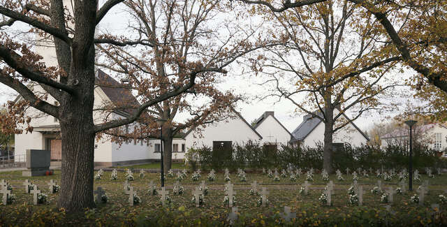 A graveyard in front of a white church. Small stone crosses can be seen in multiple rows.