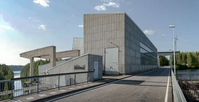 Hydropower plan seen from the bridge that spans across the river. The main facade materials are concrete and metal. Four rows of ribbon windows decorate the facade. The architecture could be describes as rather industrial and Functionalistic.