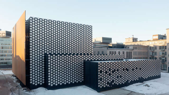 Wall of large cube shaped building made of small round metal shapes. Wall is in three levels. Blue sky in background.