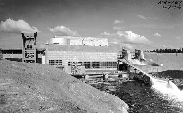 An old black and white photograph from the river. The hydropower plant is in use here, and water is flowing through the machinery.