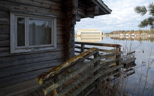 In front a wooden, log-built cottage by the lake. In the background on the other side of the lake lies the power plant.