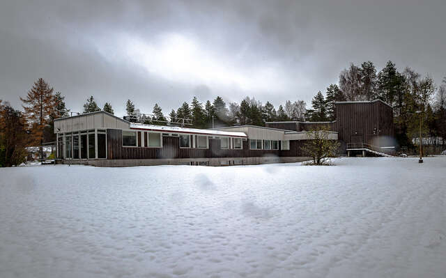 A building in a snowy landscape with a façade that consists of vertically layed, dark wooden blanks and windows decorated with white wooden frames.