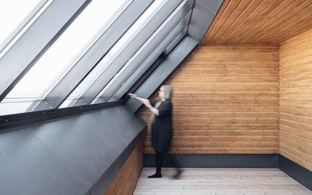 A room with a row of widows tilted inwards give light to the wooden ceiling and walls. One person is standing by the windows.