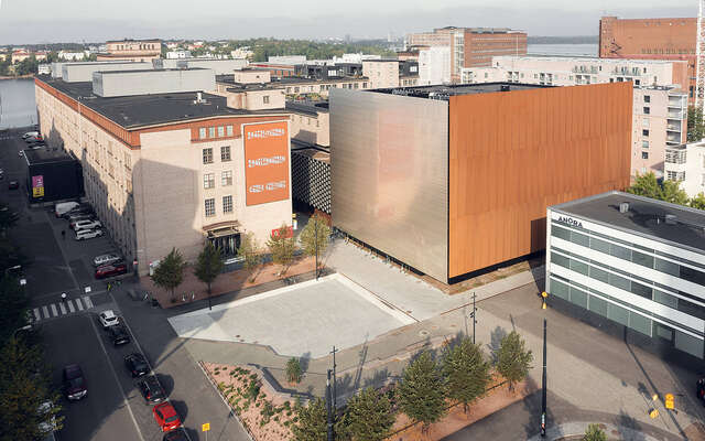 Aerial view of buildings with large cube shaped building in center with one metal colored wall and one bronze colored wall.