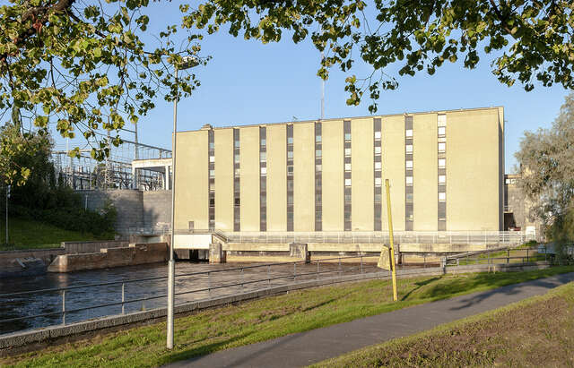 Photo taken from the shores of the river, showing the river and the façade if the power plant. The facade has light stone. A darker shade is used in the shape of a column to highlight high vertical windows that stretch from the ground to the roof, covering the whole façade.
