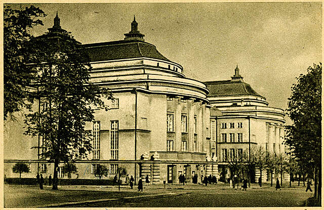 Old black and white picture of a grand building with people walking in the square in front of it.