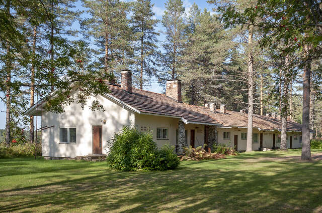 Photo showing a house plastered with a natural white shaded plaster, and with a roof made of red roof tiles, as well as chimneys that are laid with red bricks. The image also shows that the details of the window frames and doors were built out of wood.