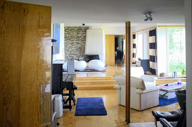 A photo showcasing the living room, that is divided into two levels, the level in the front of the image being the lower area, and the area at the back of the image being the area lifted with a couple of steps. This makes the space more interesting, creating an architectural hierarchy in the big, open nature of the living room. Light flows into the space from the high windows on the right side of the image. The wall colour is white and the the floor flooring is shiny, treated wood flooring. At the back of the image the space is being defined by a strong architectural element that is the white plastered fireplace, combined with the adjacent dark brick wall decoration.