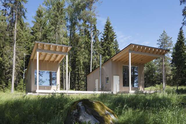 Two square-shaped wooden huts are next to each other, surrounded by forest.