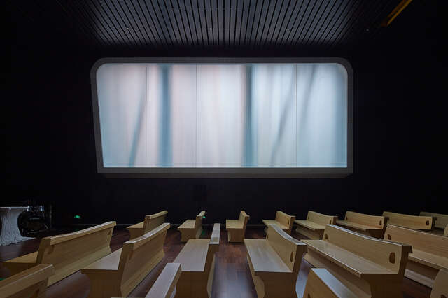Interior with light-coloured wooden benches and a large window pointing out from a dark background