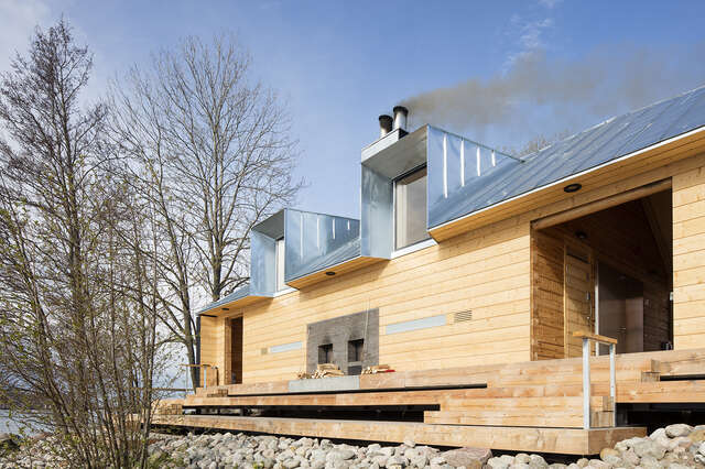 Wooden sauna building with a handcrafted wooden log structure left bare and a sculptural pitched roof in zinc plate.