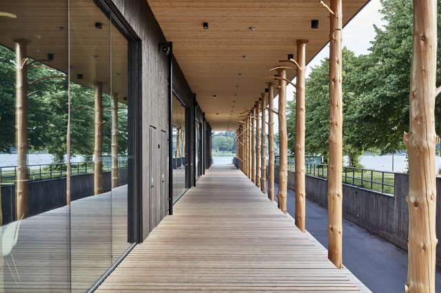 Long wooden terrace with wooden pillars on the right and large glass windows on the left.