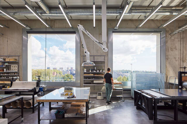 Classroom with two large square windows and person standing between the windows. Large tables can be seen in the room, as well as a large withe lamp hanging from the ceiling.