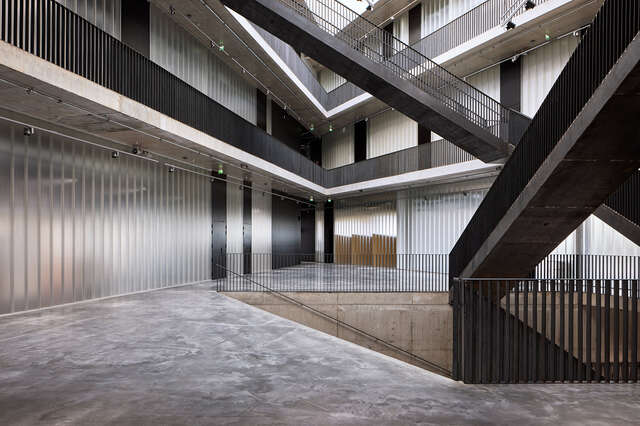 Interior of concrete structure. Light coloured wall with black doors in the centre of the image, large black staircases on the right.