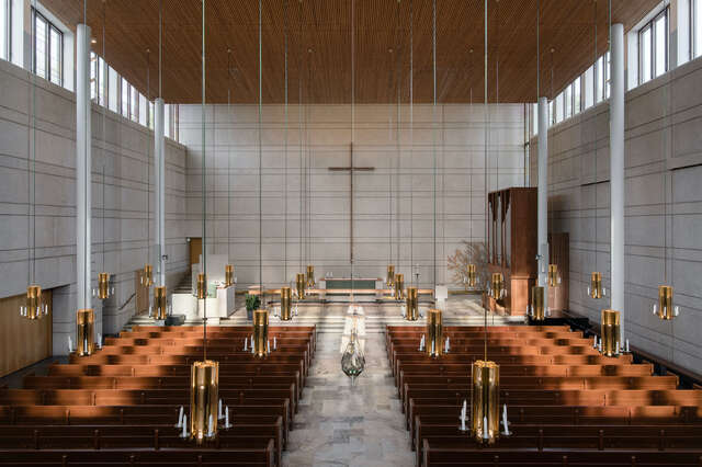 A high space with a band of windows below the ceiling. Rows of benches, cross on the wall, lapms hanging.
