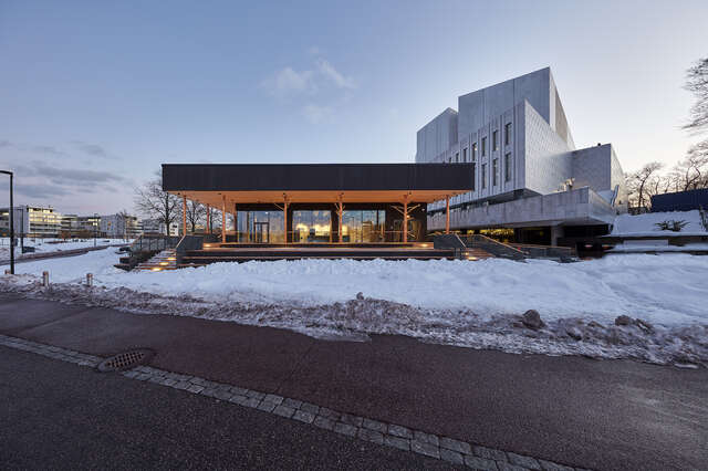 A low dark wooden building with lighting next to a larger white structure. Asphalt and snow in the foreground.