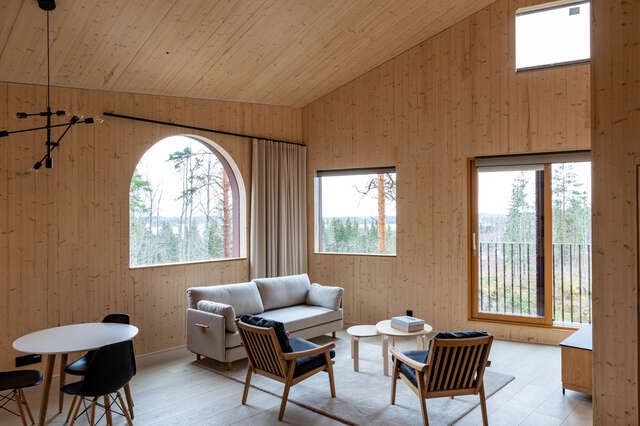Modern room with smooth, untreated wooden walls and ceiling, several windows, sofa and chairs