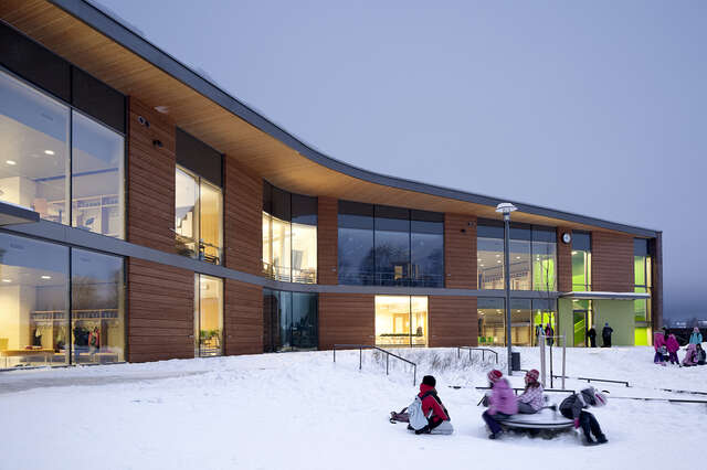 Snowy school yard with children playing outside, in the background the wooden school building is illuminated s the sun is beginning to set.
