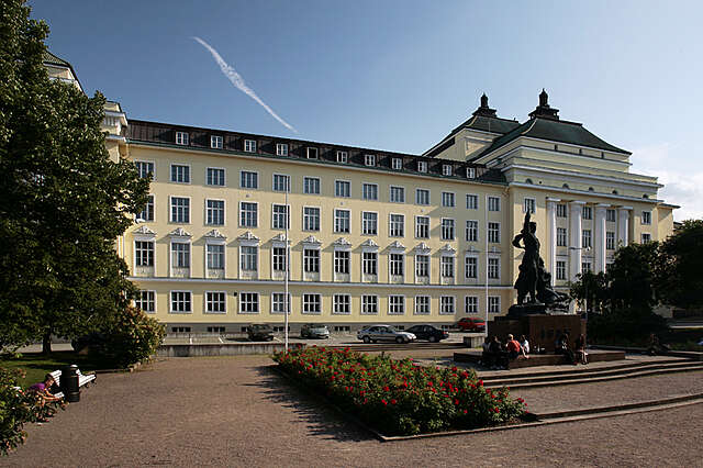 Yellow multiple storey building with white structural elements and decorations and a park in front of it.