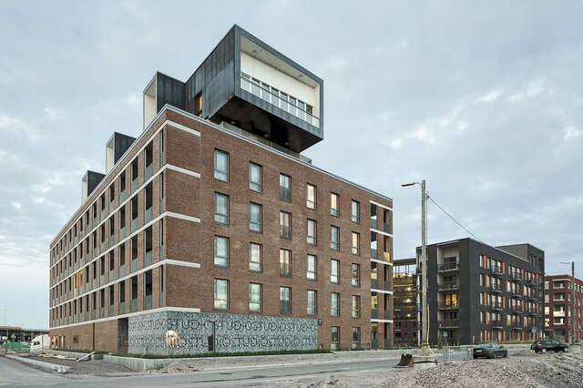 Red-Brick facade of te building with a black and white extension on the top, viewed from the other side of the street.