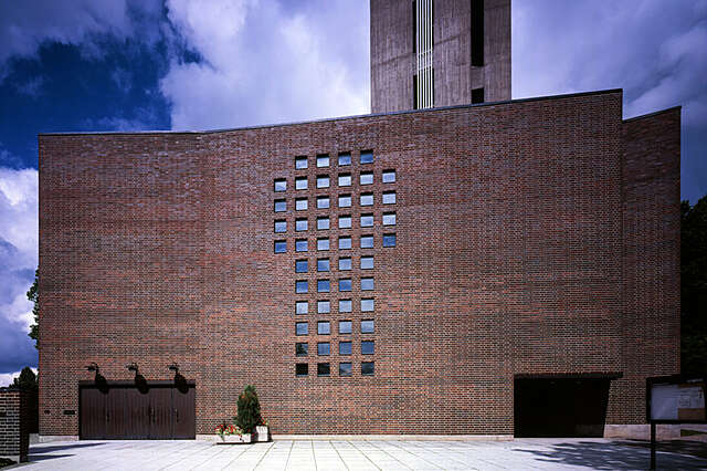 Red brick church with a glass detail at the front and a massive bell tower of raw concrete, integrated into the northwest corner of the church.