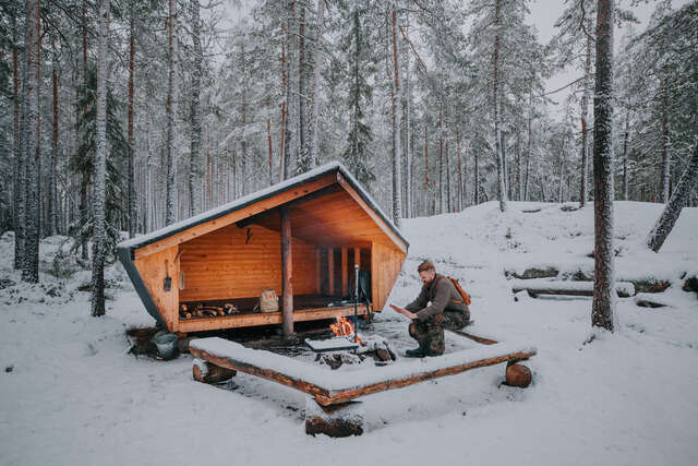 A wooden structure in a snowy forest. Small campfire with on person sitting in foreground.