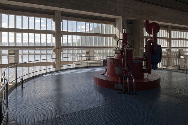 Photo of the machine hall with bright red machinery on oval-shaped, blue floor. Natural light flows into the space from large windows in the background of the image.