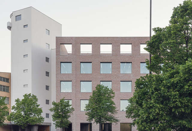 Large brick building as seen from outside. 4 green trees are in front of the building
