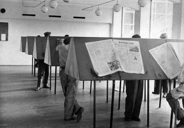 Triangular reading tables with people standing between them and reading newspapers in a white environment with circular lamps