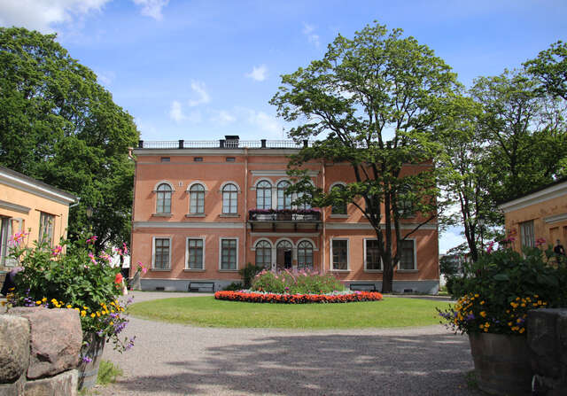 Terracotta coloured facade with grid windows and arch details. In the front there is a flowerbed and a tree.