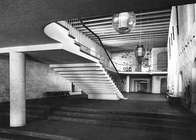 Black and white picture of a hall in an old stone building with white stairs leading to the second level balcony.