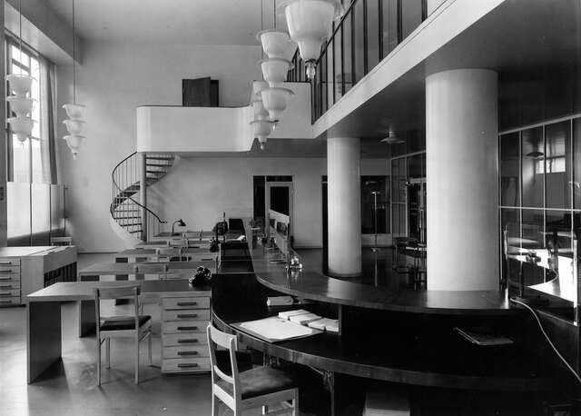Black and white picture of an office with a curved reception desk and several smaller office desks next to it. At the back of the room there is a spiral staircase leading in to the second floor.