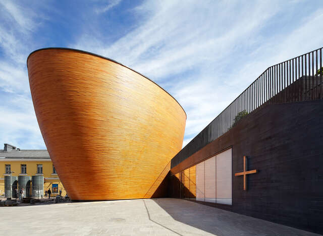 A wooden sculptural, curved box connected to a lower, rectangular part with a cross symbol on the façade