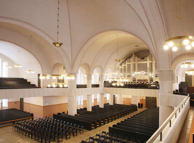 Church interior with a view towards the organ. The mostly white interior has terra-cotta and black elements and tall arched ceilings.