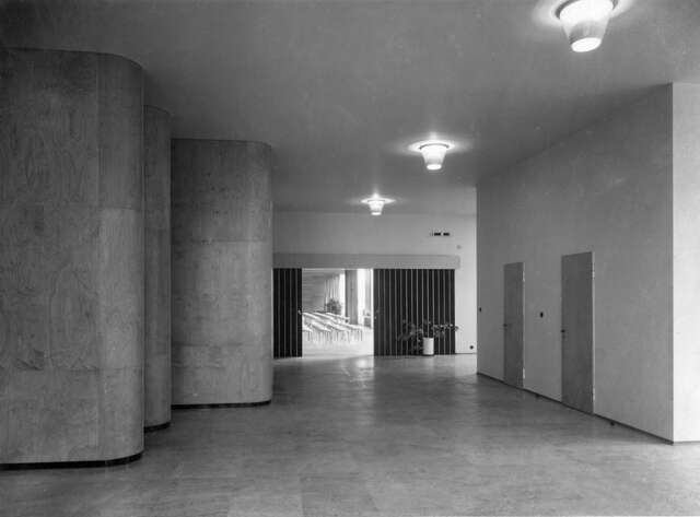 Light interior with oval shaped wooden pillars in the middle of the room, a auditorium is visible behind black sliding doors.
