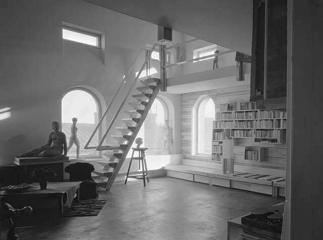 Black and white image of a light wooden interior with arch windows, female sculptures and a bookshelf with a wooden ladder leading upstairs.